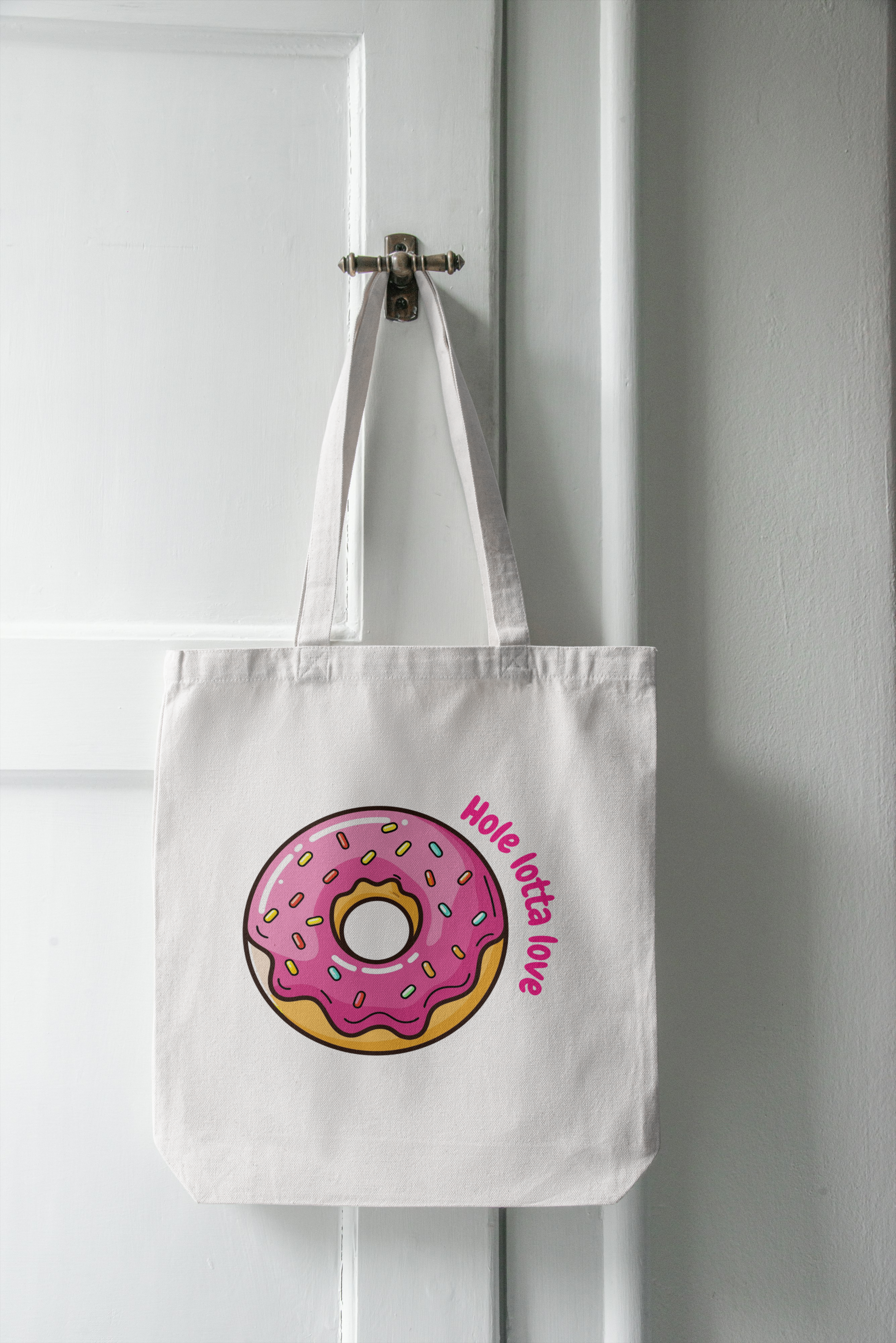 White tote bag with pink donut design and 'Hole lotta love' text hanging on a white door.
