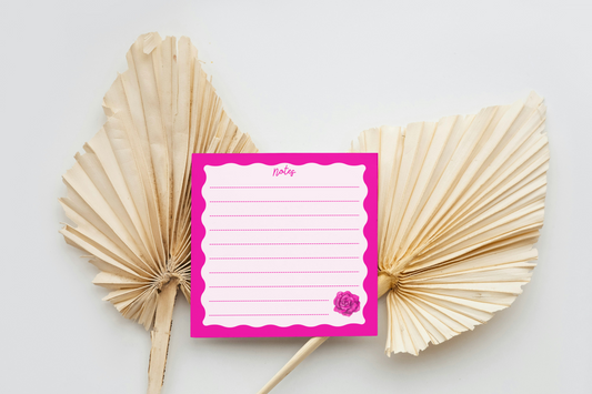 Memo pad with pink cover and white pages between two dried palm leaves on a light background