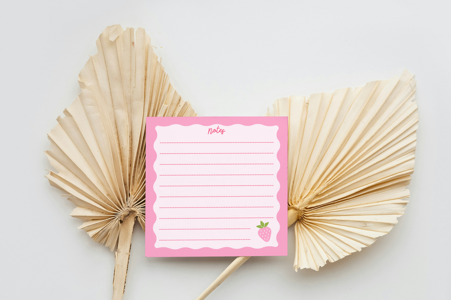 Pink memo pad with a strawberry design between two dried leaf fans on a light background