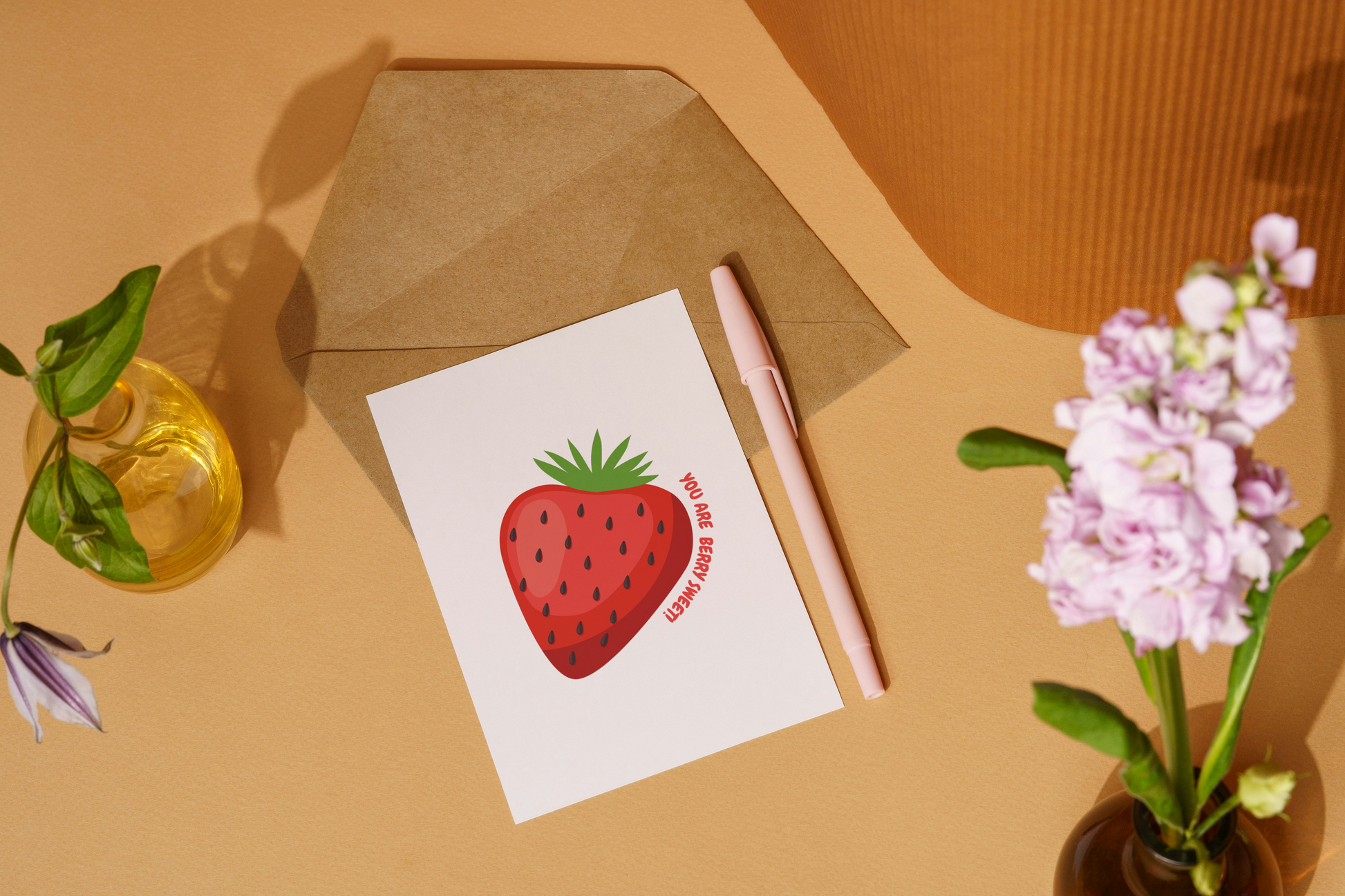 Card with strawberry illustration on a beige surface with pen and flowers