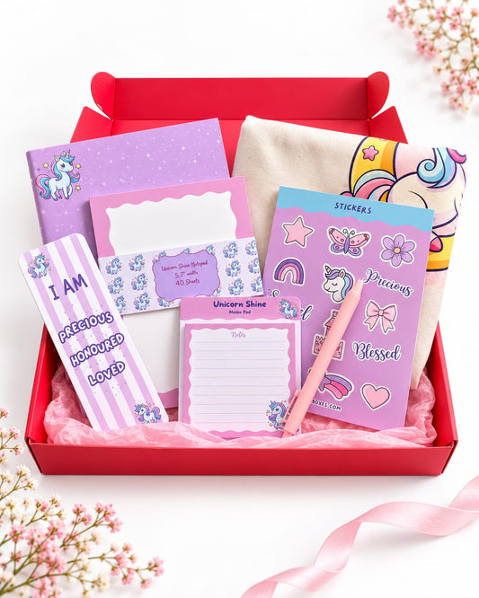 A red gift box filled with pastel unicorn-themed stationery and accessories, including a notepad, a pink pen, stickers, and a tote bag, arranged on a white background with pink ribbon and baby's breath flowers.