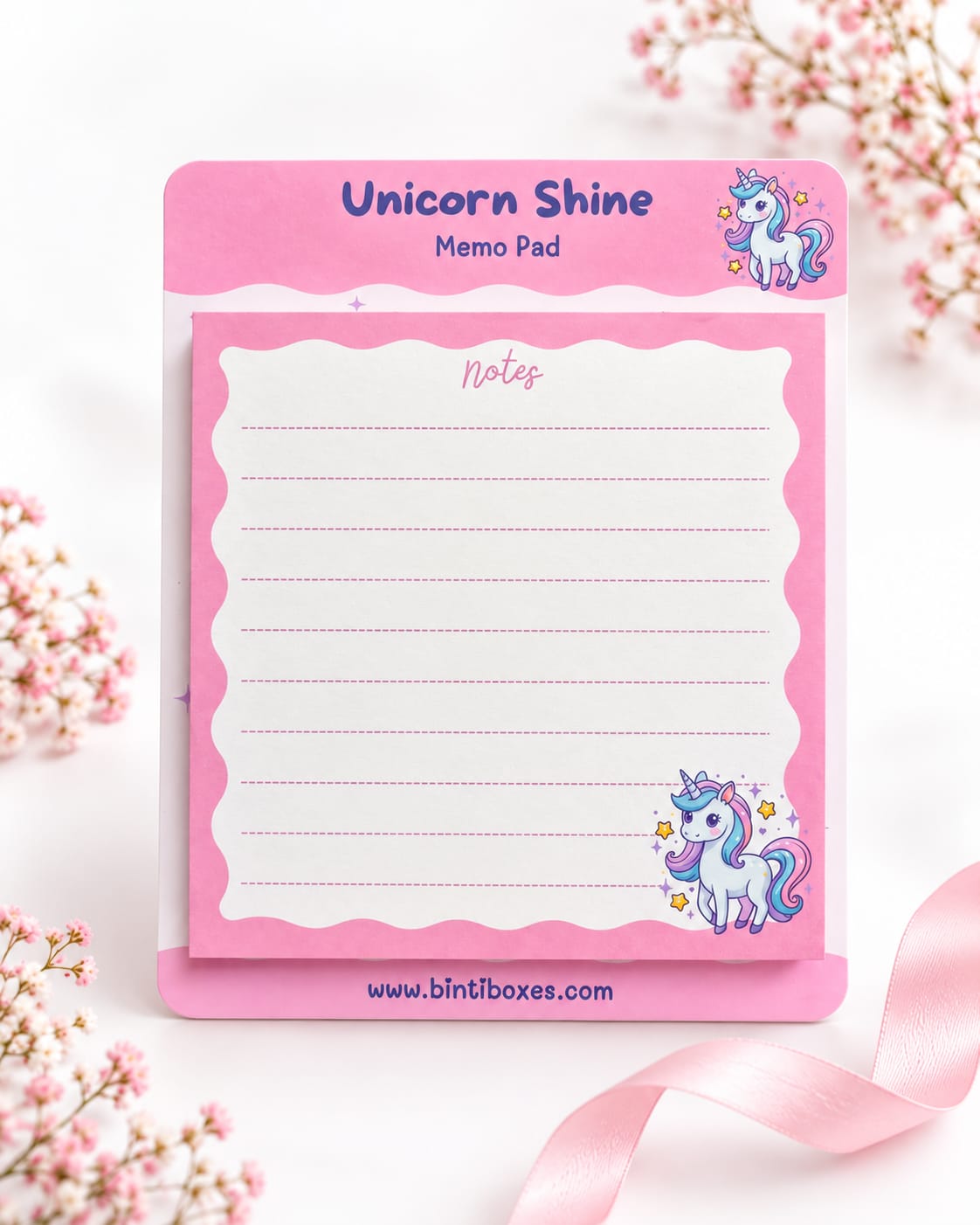 A pink memo pad with unicorn illustrations, labeled 'Unicorn Shine Memo Pad,' photographed on a white surface with soft pink flowers and a ribbon around it.